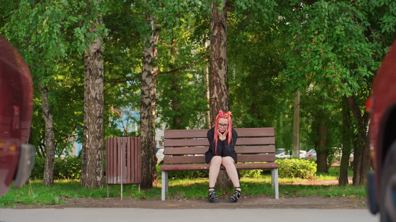 자작나무 아래 벤치에 앉아 차 사이에서 기다리는 백인 학생, 전경을 둘러싼 빨간색 차들, 백팩과 헤드폰, 발에는 스케이트, 불안하고 외로운 분위기, 햇빛이 비치는 여름 잎, 도시 길가