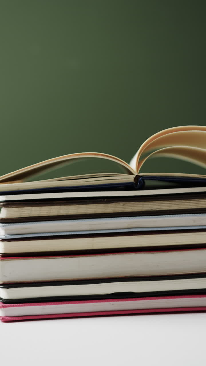 Vertical video of open book on stack of books with copy space on green background, in slow motion