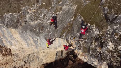 Escaladores-Colgando-De-Cuerdas-Desde-El-Borde-De-Una-Montaña-Rocosa-Perforando-Las-Rocas-Con-Un-Dron-Disparado