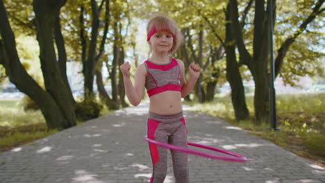 Athletic-fitness-toddler-girl-training-playing-twisting-Hula-hoop-circle-ring-around-waist-in-park