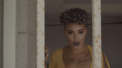 witchy, tattooed woman wearing a yellow dress walking behind rusty white bars