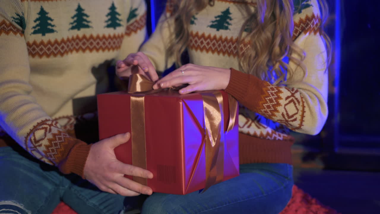 Couple in Christmas sweaters with a box gift. Woman unties ribbon from the present box. Romantic atmosphere during New year celebration. Close-up.