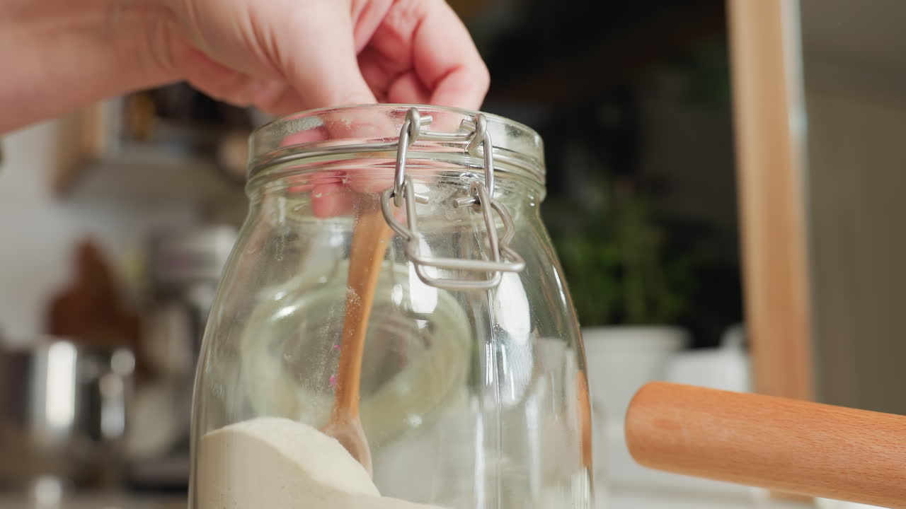 Hand view of chef scooping corn flour with wooden spoon from glass jar, gently shaking to level for accurate measurement, with visible dust particles rising in warm kitchen
