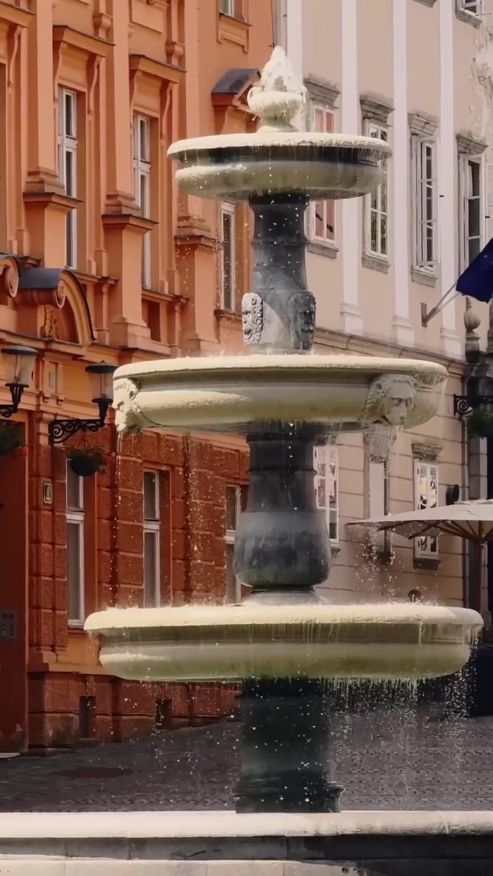 Vertikalaufnahme des Novi Trg Brunnens, der vor historischen Fassaden im Zentrum Ljubljanas herabstürzt, wobei slowenische und EU-Flaggen der Altstadtszene einen bürgerlichen Charakter verleihen.