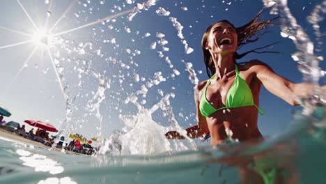 low-angle video shot of a joyful woman in a green bikini splashing water at the beach