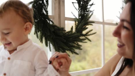 Young-mother-is-holding-her-son's-hands-while-he-is-walking-on-the-window-sill-decorated-with-Christmas-wreath.-Happy-family