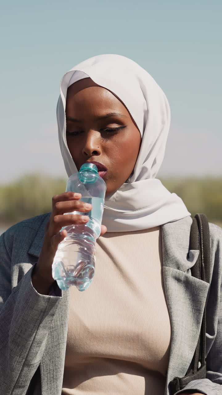 Tired thirsty Muslim woman opens bottle of water and drinks at riverside on hot sunny day. Lady with hijab refreshes during promenade slow motion