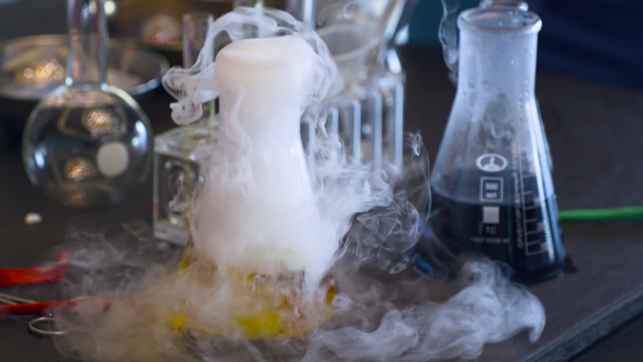Primo piano della drammatica sublimazione di ghiaccio secco che crea una fitta nebbia bianca che straripa da un liquido giallo in un pallone di Erlenmeyer, circondato da vetreria e strumenti da laboratorio in un allestimento per esperimenti scientifici.