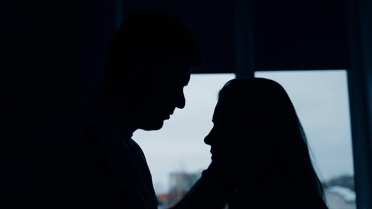 Silhouettes of a couple in love in a dark room standing in front of window. Man soothing and kissing his woman.