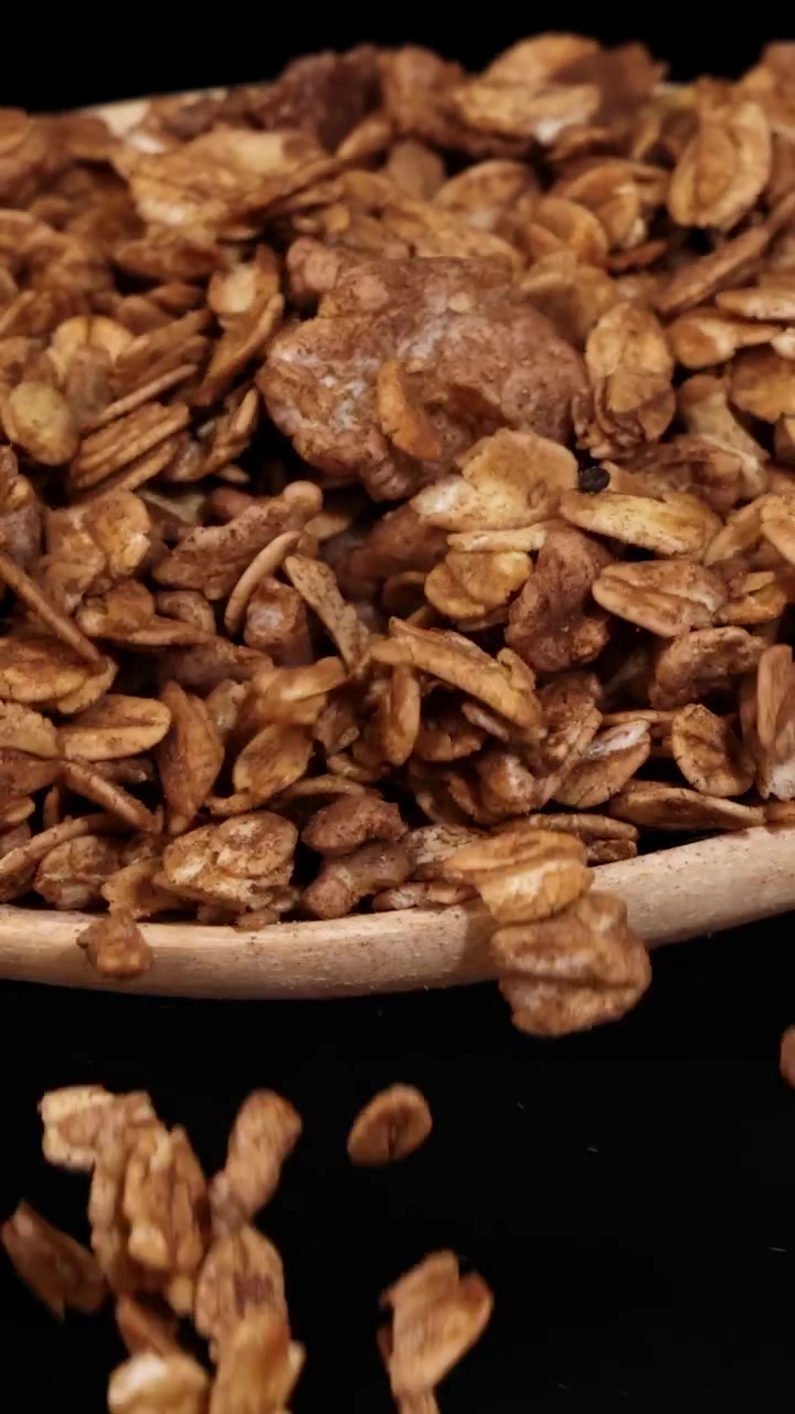 Pezzi di granola cadono a cascata da un cucchiaio, ripresi al rallentatore su uno sfondo scuro, evidenziando la consistenza e il movimento.