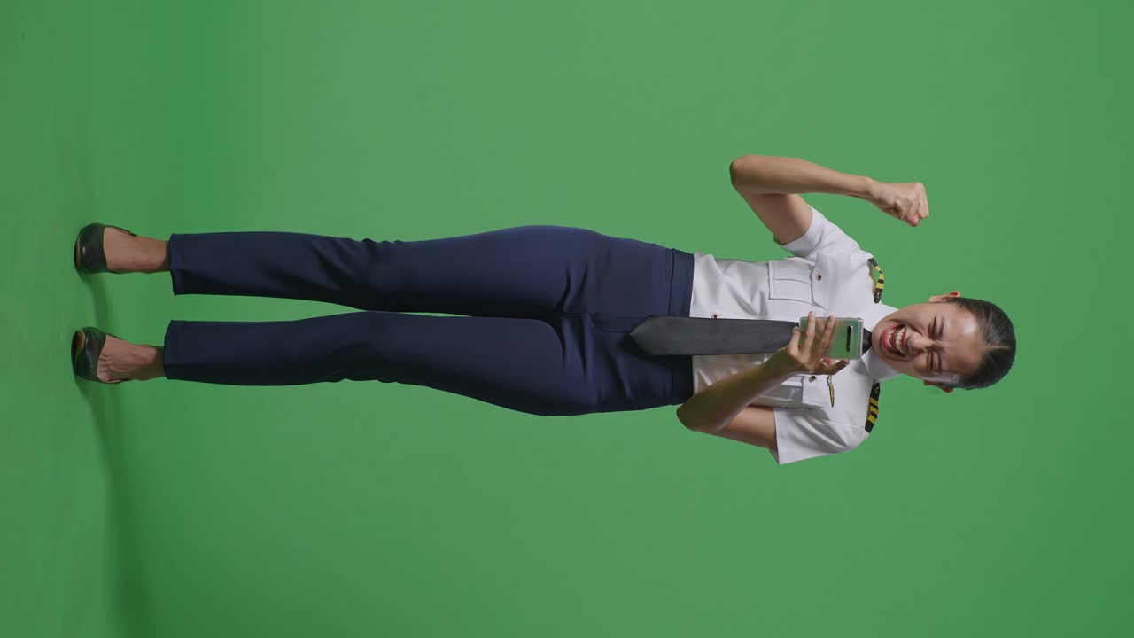 아시아 여성 조종사가 스마트폰을 보며 소리를 지르며 골을 축하하고 녹색 스크린 배경 스튜디오에 서 있습니다.