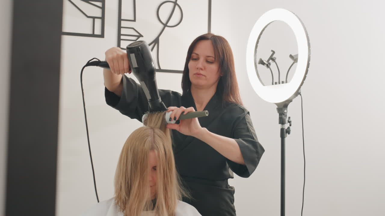 blonde client salon blowdry under ringlight, focused stylist uses round brush and hot air to smooth damp hair, modern wall art and soft studio lighting create precise, professional atmosphere