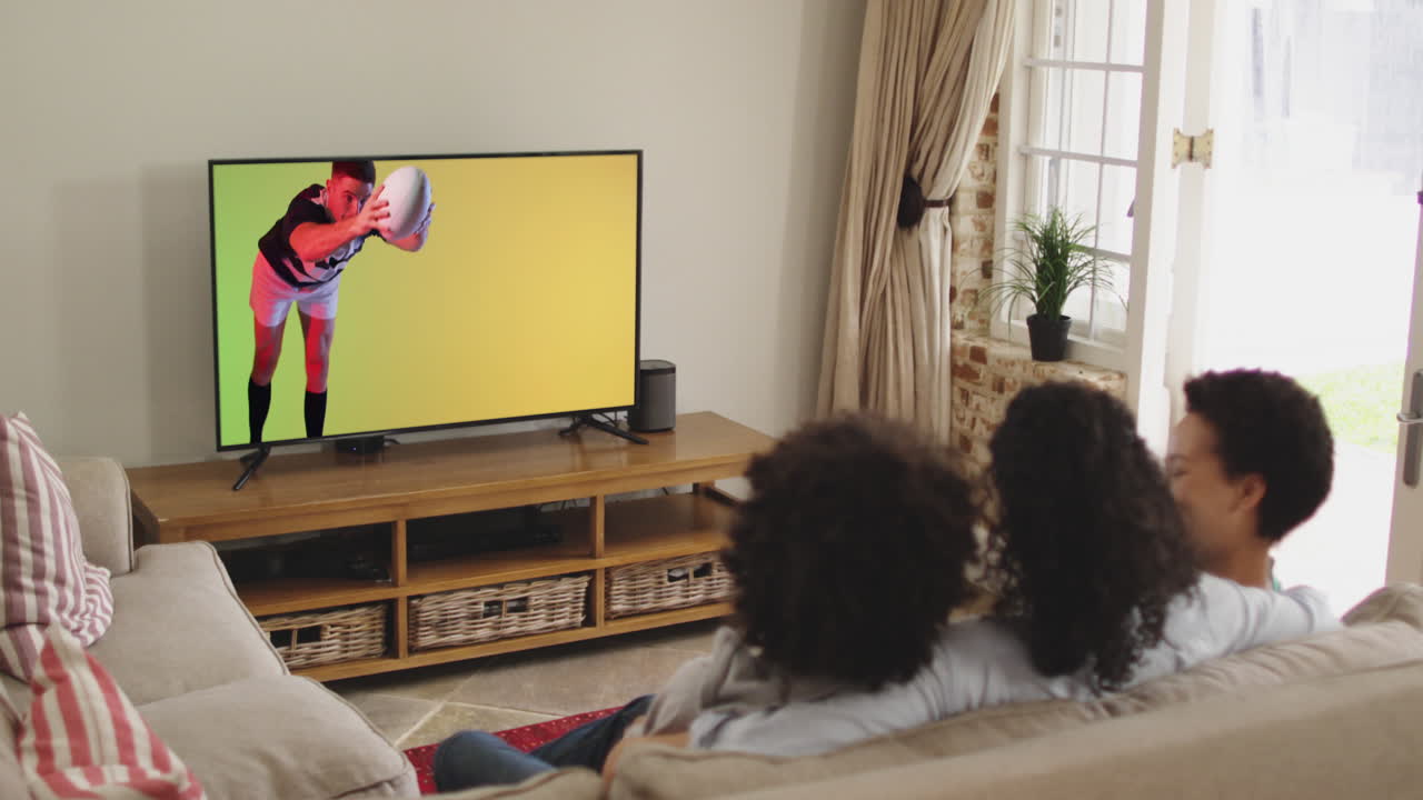 familia biracial viendo la televisión con un jugador de rugby caucásico saltando con la pelota en la pantalla