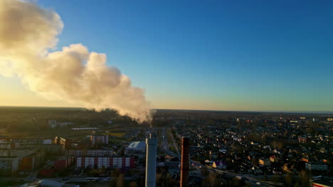 Cierre-De-Paralaje-Aéreo-Con-Torre-De-Fábrica-Industrial-En-Primer-Plano,-Puesta-De-Sol-Vívida-A-Través-Del-Humo