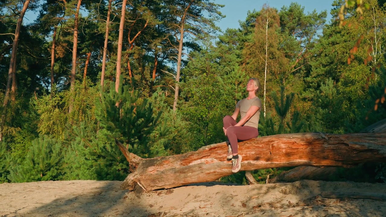 Caucasian woman sits peacefully on a tree trunk enjoying warm sunshine, with a lush forest behind her beneath a clear blue summer sky.