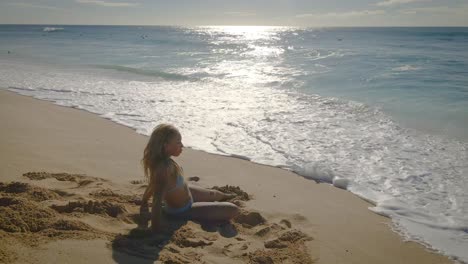 with yellow sand beneath her, the girl in the blue bikini basks in the beauty of the turquoise water waves that stretch out before her on the tropical beach