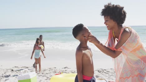 familia afroamericana sonriente usando crema solar en una playa soleada