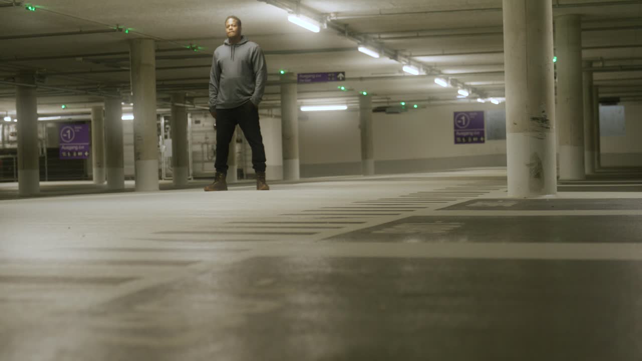 View of cool black male standing with hands in pocket inside empty underground parking lot with bright lights in Metzingen, Germany, Baden Wurttemberg, Europe, panning view angle