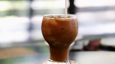 una mano agita el café helado con una pajita, destacando patrones giratorios y condensación en un entorno brillante y relajado
