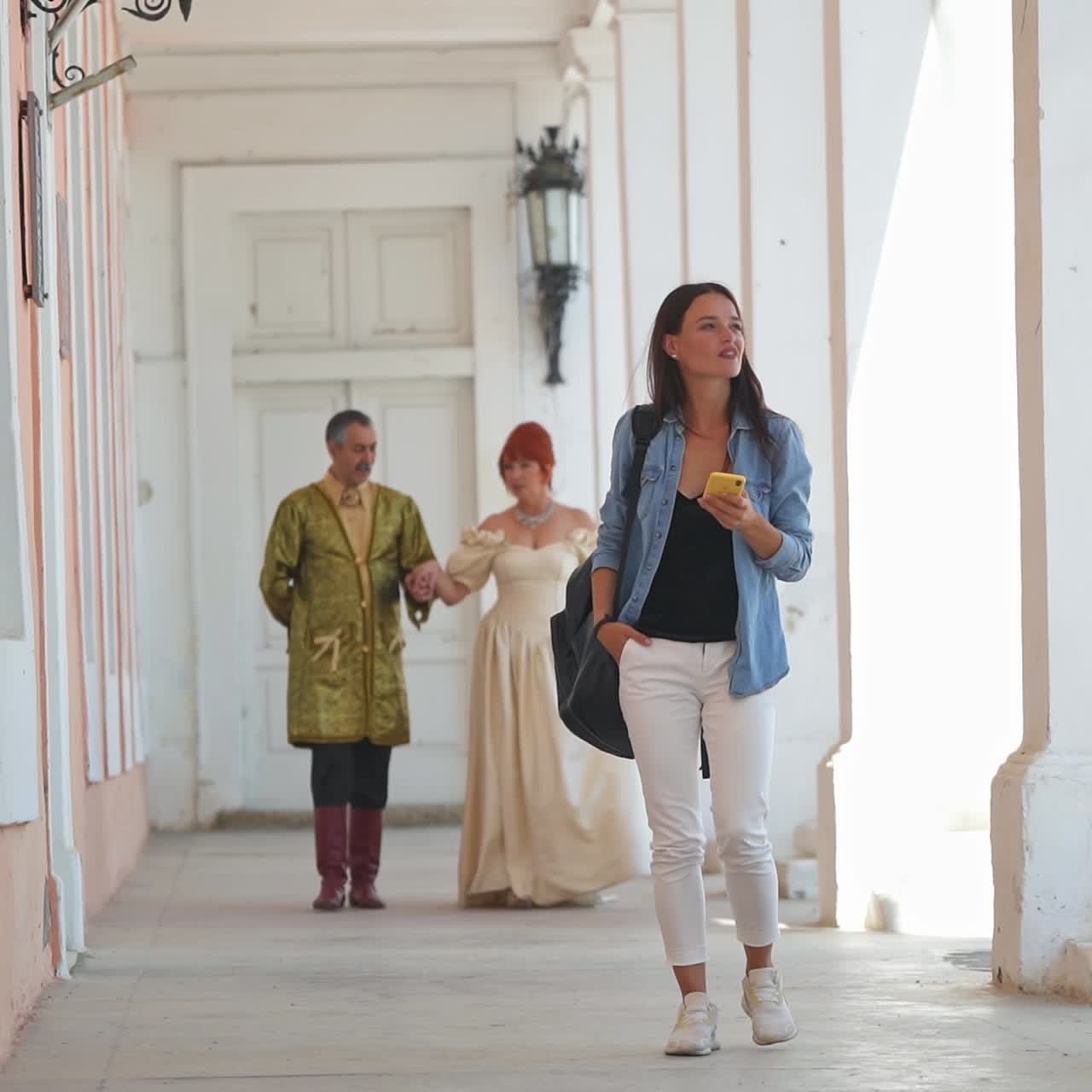 Adult couple in old-fashioned clothes walking by the long hall holding by hands. Girl in modern look appears taking selfie. Present meets past