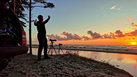 hombre emocionado al lado de un trípode con cámaras instaladas mirando a la impresionante vista de la orilla del océano desde la playa exclamando emoción al atardecer