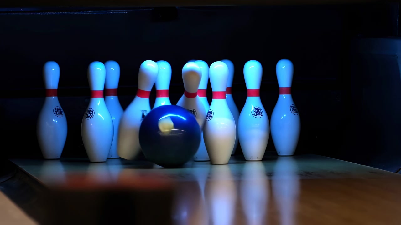 Pohled z nízkého úhlu na dráhu ukazuje černou bowlingovou kouli, jak se kutálí do trojúhelníkového stojanu deseti bílých kuželek s červenými pruhy, spouštějící náraz, rozhazující kuželky, lesklé odrazy a dynamickou sportovní energii.