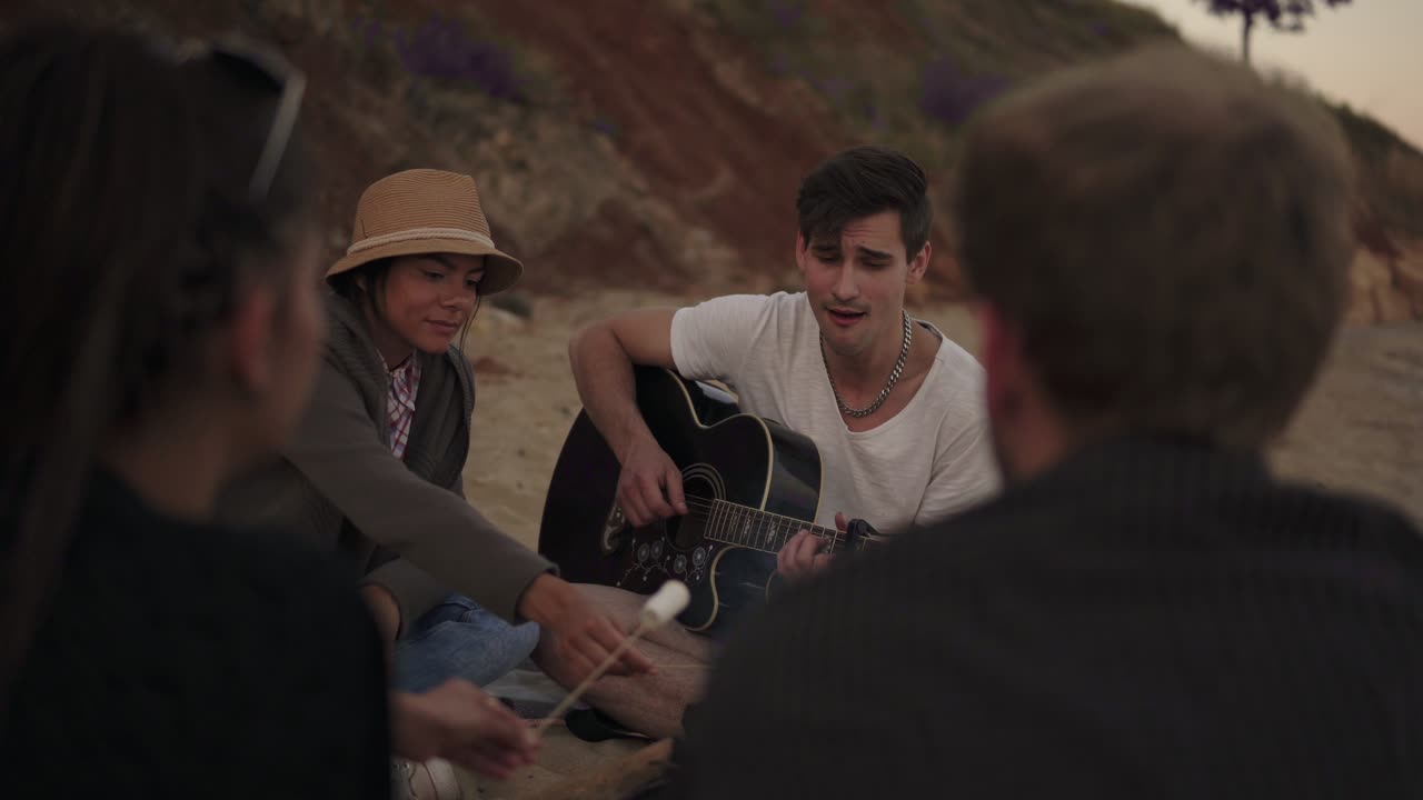 Young cheerful friends sitting by the fire on the beach in the evening, cooking marshmallow and playing guitar. Shot in 4k