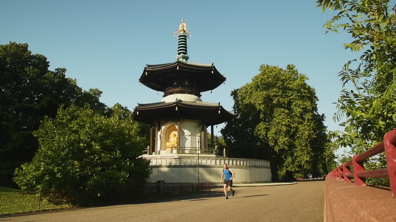 A fit red-haired man run in a blue shirt on the dirt paths past the Battersea pagoda in London during the midday sun on a blue sky day