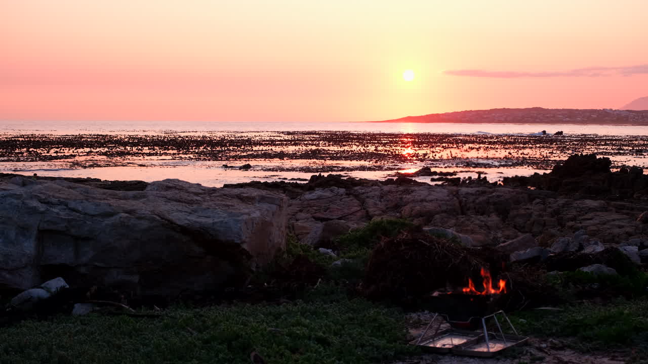 Small fire burning on portable barbecue on rocky coastline during vivid sunset
