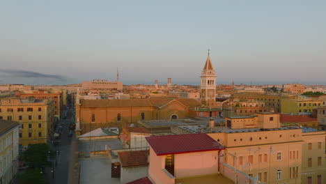 Forwards-fly-above-buildings-in-city-centre,-old-church-with-tower-lit-by-morning-sunshine.-Rome,-Italy