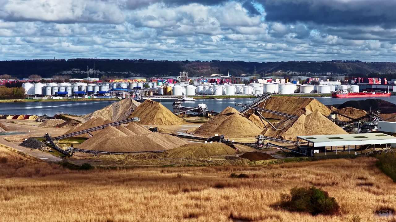 Industrial port storage tanks conveyors and sand piles beside harbor in Le Havre France under cloudy sky, aerial establishing orbit