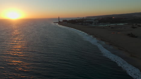 Aerial-view-in-orbit-over-the-shore-of-Maspalomas-beach-during-sunset,-with-the-Maspalomas-lighthouse-and-the-pond-in-the-background