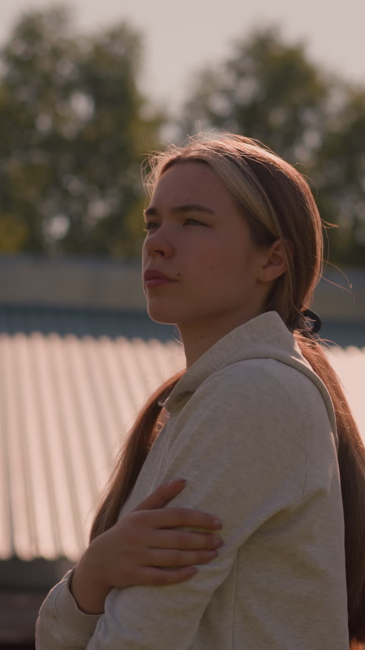 una joven con el cabello largo atado hacia atrás en una sudadera con capucha se encuentra al aire libre, mirando pensativa con expresión reflexiva, la luz del sol arroja un brillo cálido sobre ella, con el techo y los árboles en el fondo