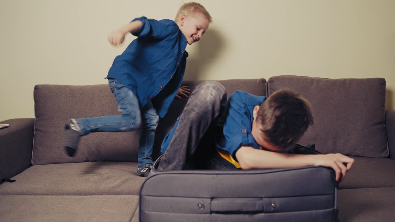 Two brothers are playing on the couch with travelling bag in the living-room. Elder boy is trying to lay himself into a grey suitcase at home.