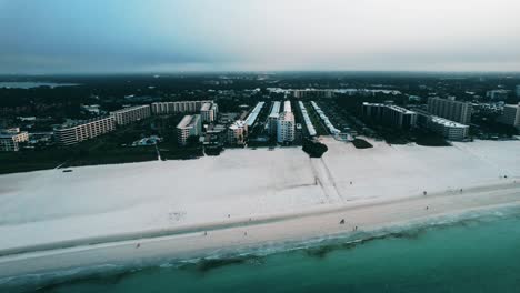 Drone-clip-following-shoreline-between-clear-blue-green-sea-and-white-sand-beach,-with-cityscape-stretching-to-horizon