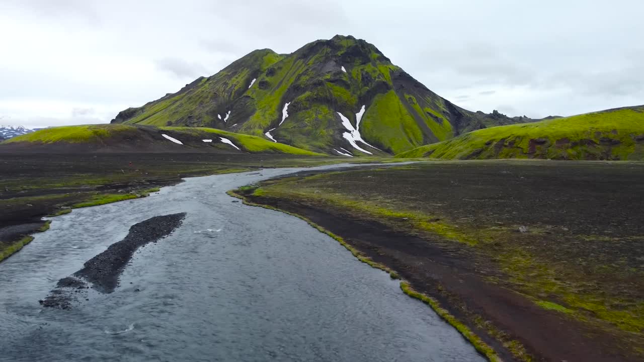 Aerial drone footage flying over a cold fast flowing small river flowing in Iceland during cloudy weather day, with large mossy and green steep snow covered mountain in the background. Moss visible.