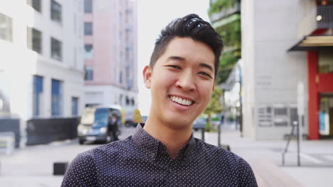 Millennial-Asian-man-sitting-in-a-city-street-laughing-to-camera,-head-and-shoulders-close-up