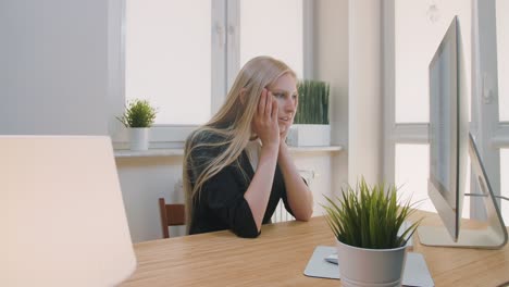 upset woman sitting at workplace. blond sad female in elegant clothes clutching head after failure or bad news sitting in light office at desk with computer.