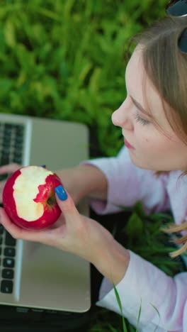 programadora acostada en la hierba en un parque trabajando en una computadora portátil con una manzana en la mano, tomando un bocado mientras sonríe, rodeada de exuberante vegetación, luz natural destacando su enfoque en el entorno de trabajo al aire libre