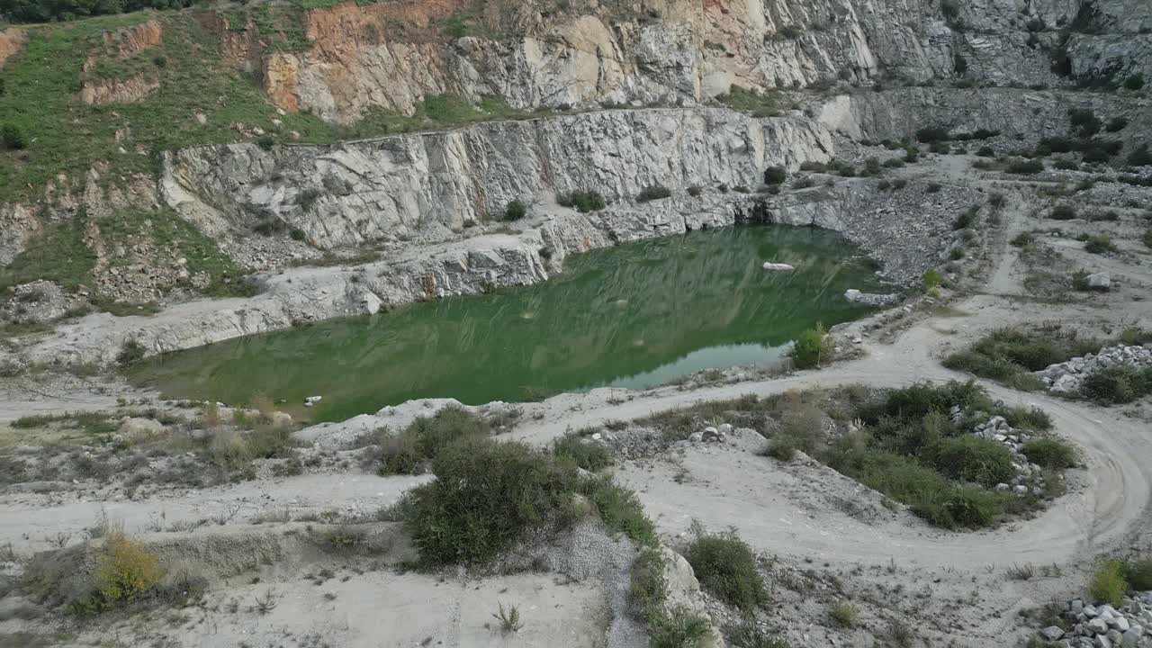 Green hued quarry pit nestling between rocky cliffs, revealing scarred landscape with sparse vegetation under aerial perspective, showcasing industrial mining terrain and environmental impact