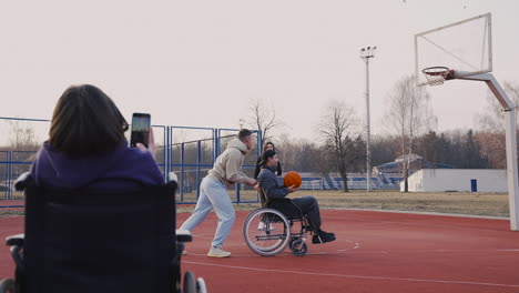 mujer discapacitada en silla de ruedas grabando con smartphone a sus amigos jugando al baloncesto 2