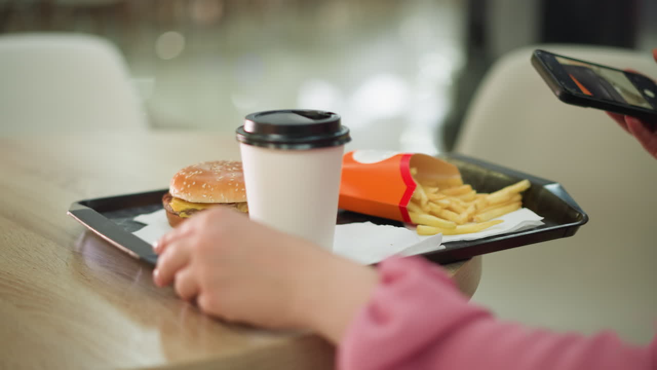 una mujer ajusta una bandeja negra en una mesa de madera que contiene una taza de café, una hamburguesa y patatas fritas con la mano izquierda mientras sostiene su teléfono en la mano derecha, el fondo está suavemente borroso