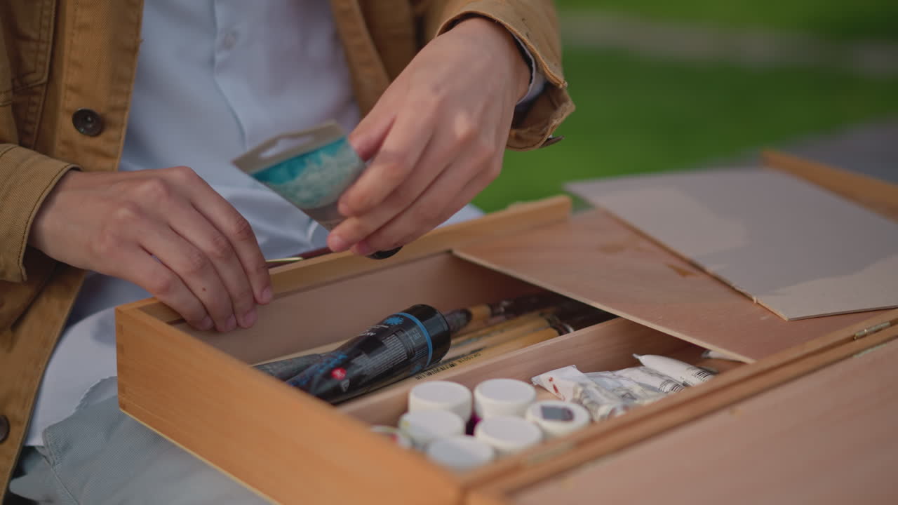 Hvid kunstner arrangerer trækasse med maling på bænk og udvælger tuber, paletkniv og pensler. Organiserer rum, lukker haspen. Blødt, naturligt lys, antydning af skitsebog med gadekunst, rolig og eftertænksom