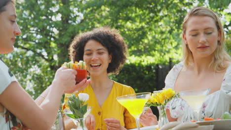 three female friends sitting outdoors in summer garden at home drinking cocktails and eating meal