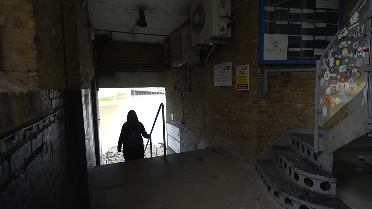 Woman walks down old stone stairs toward the Thames River in a moody, urban passageway