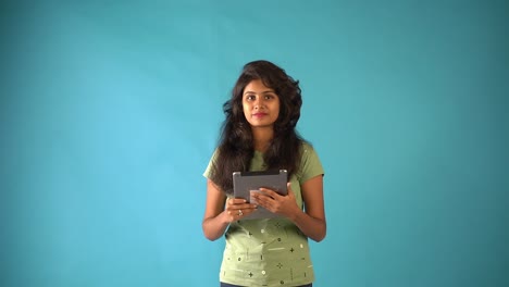 A-young-Indian-girl-in-orange-frock-standing-with-a-tab-looking-at-the-camera-in-an-isolated-blue-background-studio