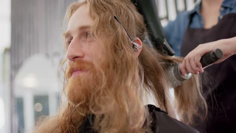 Caucasian-female-hairdresser-styling-male-client's-long-hair-with-hairdryer-and-brush,-slow-motion