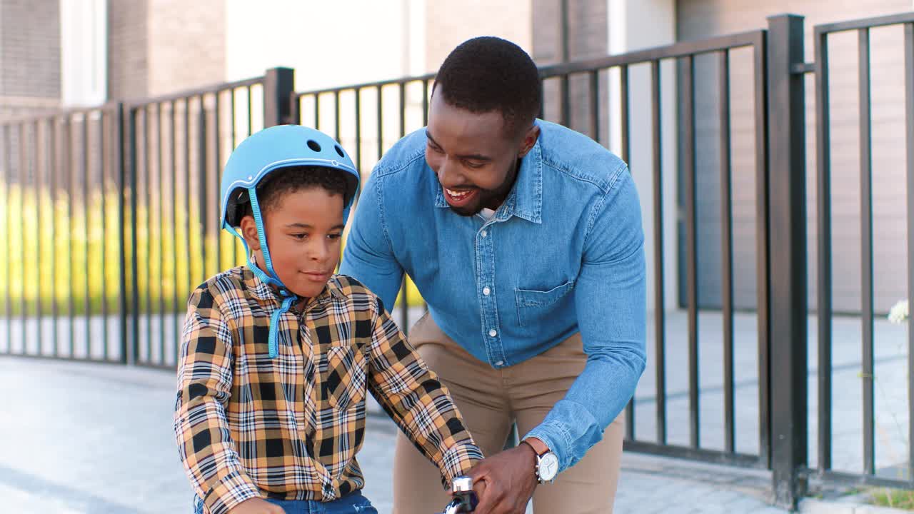 feliz padre afroamericano enseñando a un niño pequeño con casco montando en bicicleta en la calle en las afueras