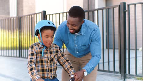 Glücklicher-Afrikanisch-amerikanischer-Vater-Unterrichtet-Kleinen-Jungen-Im-Helm-Beim-Fahrradfahren-Auf-Der-Straße-Am-Stadtrand