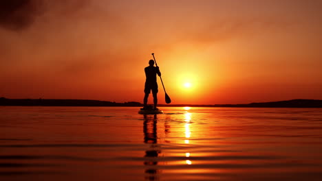 dark male silhouette standing on sup board on the river. man rowing slowly distancing from the camera.